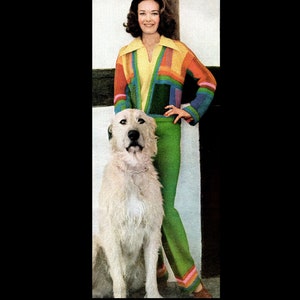 Può includere: Una donna indossa un maglione colorato con motivi geometrici e pantaloni verdi e sta accanto a un cane bianco. Il maglione ha un colletto giallo e un motivo verde, blu e viola.