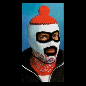 Puede incluir: Un pasamontañas tejido en blanco, rojo y negro. La máscara presenta un pompón rojo en la parte superior, aberturas negras para los ojos y la boca, y un calentador de cuello estampado en rojo y blanco. El diseño se asemeja a una cara con bigote y barba.