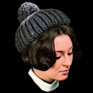 May include: A grey knitted hat with a pompom on top. The hat is being worn by a person with dark hair.
