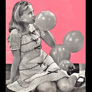 Puede incluir: Una niña pequeña con un vestido blanco de ganchillo con detalles de cinta negra sopla un globo rosa. Está rodeada de otros globos en tonos grises.