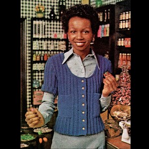 May include: A blue crocheted cardigan with a button-up front and short sleeves. The cardigan is worn over a white and blue patterned shirt.