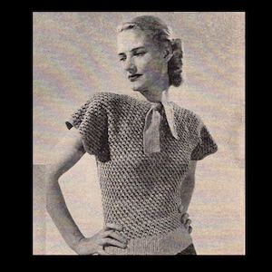 May include: A close-up of a woman wearing a knitted top with a bow at the neck. The top is a light grey colour and has a textured pattern. The woman is standing with her arms crossed and is looking to the side.