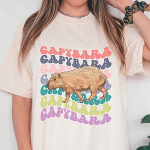 K&ouml;nnte beinhalten: Ein wei&szlig;es T-Shirt mit einem Grafikdruck eines Capybaras im Retro-Stil. Der Text "Capybara" wird in einem Regenbogen aus Farben wiederholt.