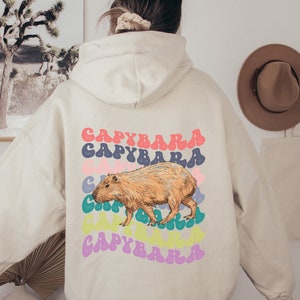 K&ouml;nnte beinhalten: Ein beiger Hoodie mit einem Grafikdruck eines Capybaras und dem Wort "Capybara", das in einem Regenbogen aus Farben wiederholt wird.