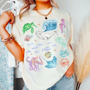 May include: A light yellow t-shirt featuring a colourful watercolour design of various sea creatures, including a whale, octopus, seahorse, turtle, jellyfish, and fish.