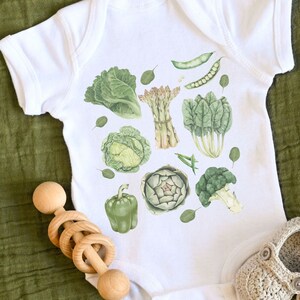 May include: White baby onesie with a green vegetable print. The print includes asparagus, artichoke, broccoli, spinach, cabbage, and bell peppers.