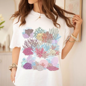 May include: White t-shirt featuring a colourful coral reef design. The design includes various types of coral, sea anemones, and fish in shades of pink, purple, blue, and yellow.