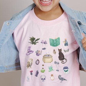 May include: A pink t-shirt featuring a collection of whimsical watercolor illustrations of magical items, including a cauldron, owl, black cat, and potion bottles. The shirt is worn by a young child, paired with a light blue denim jacket and a pink headband.