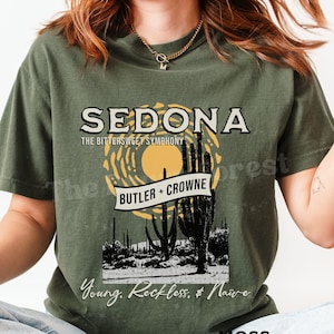 Peut inclure: T-shirt vert mousse avec un motif graphique. Le motif comprend le mot "SEDONA" au-dessus d'un paysage désertique avec des cactus et le texte "BUTLER + CROWNE". Le t-shirt comporte également le texte "Young, Reckless, & Naive".