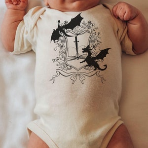 May include: A white baby onesie with a black and white graphic of a shield with a sword, book, and two dragons.