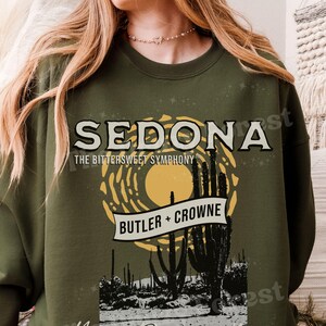 Peut inclure: Sweat-shirt vert olive avec le mot "SEDONA" dans une police vintage, avec "THE BITTERSWEET SYMPHONY" en dessous. Une scène de désert avec des cactus et un soleil est représentée. Le texte "BUTLER + CROWNE" est sur une bannière. Les mots "Young, Reckless, & Naive" sont en bas.