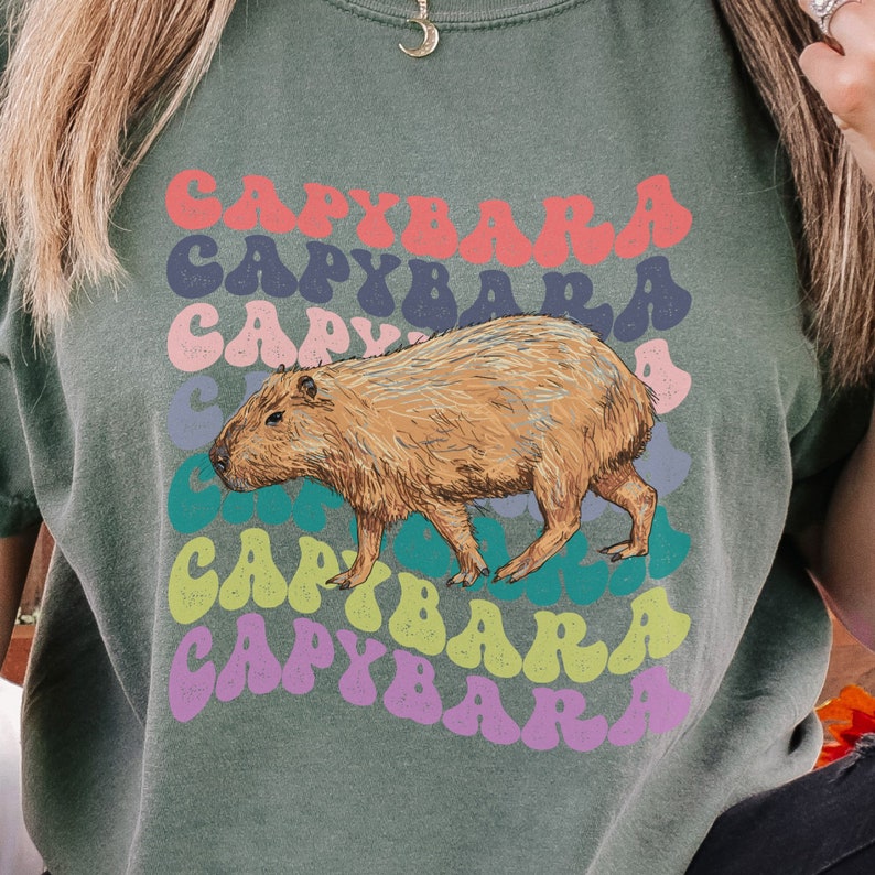 K&ouml;nnte beinhalten: Ein gr&uuml;nes T-Shirt mit einem Aufdruck eines Capybaras und dem Wort "Capybara", das in einem Regenbogen aus Farben wiederholt wird.