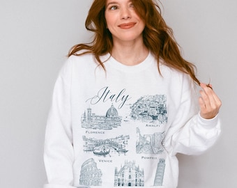 May include: White sweatshirt with a graphic print of iconic Italian landmarks, including the Colosseum, the Leaning Tower of Pisa, and the Duomo in Florence. The text "Italy" is written in cursive above the landmarks. The words "Rome", "Milan", "Pisa", "Florence", "Amalfi", "Venice", and "Pompeii" are written below the landmarks.