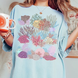 May include: A light blue t-shirt featuring a colourful coral reef design. The design includes various types of coral, sea anemones, and other marine life.
