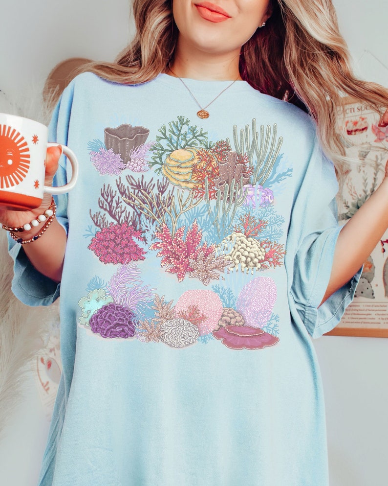 May include: A light blue t-shirt featuring a colourful coral reef design. The design includes various types of coral, sea anemones, and other marine life.