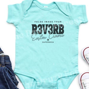 Peut inclure: Un body de bébé bleu clair avec l'inscription noire "FALSE IMAGE TOUR R3V3RB Easton Crowne SUPERNOVA". Le body a des manches courtes. Une paire de jeans bleus et des baskets blanches sont visibles.