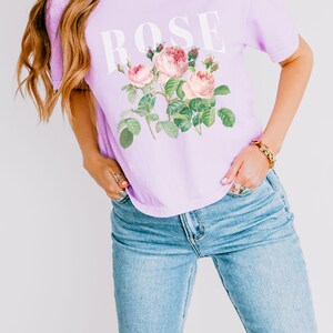 May include: A lavender t-shirt featuring a graphic of pink roses and the word "ROSE" printed in white.