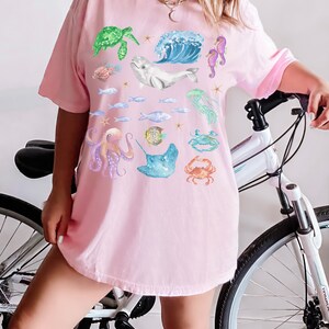 May include: A pink t-shirt featuring a colourful sea life design. The design includes a sea turtle, a whale, a seahorse, a jellyfish, an octopus, a stingray, and a crab.