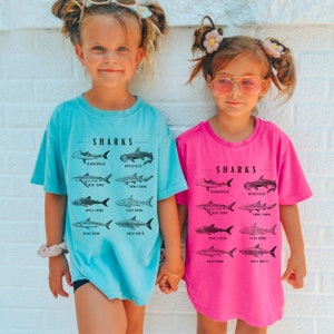 May include: Two children wearing t-shirts with a graphic of different types of sharks and the word "SHARKS" printed on them. The girl on the left is wearing a light blue shirt and the girl on the right is wearing a pink shirt.