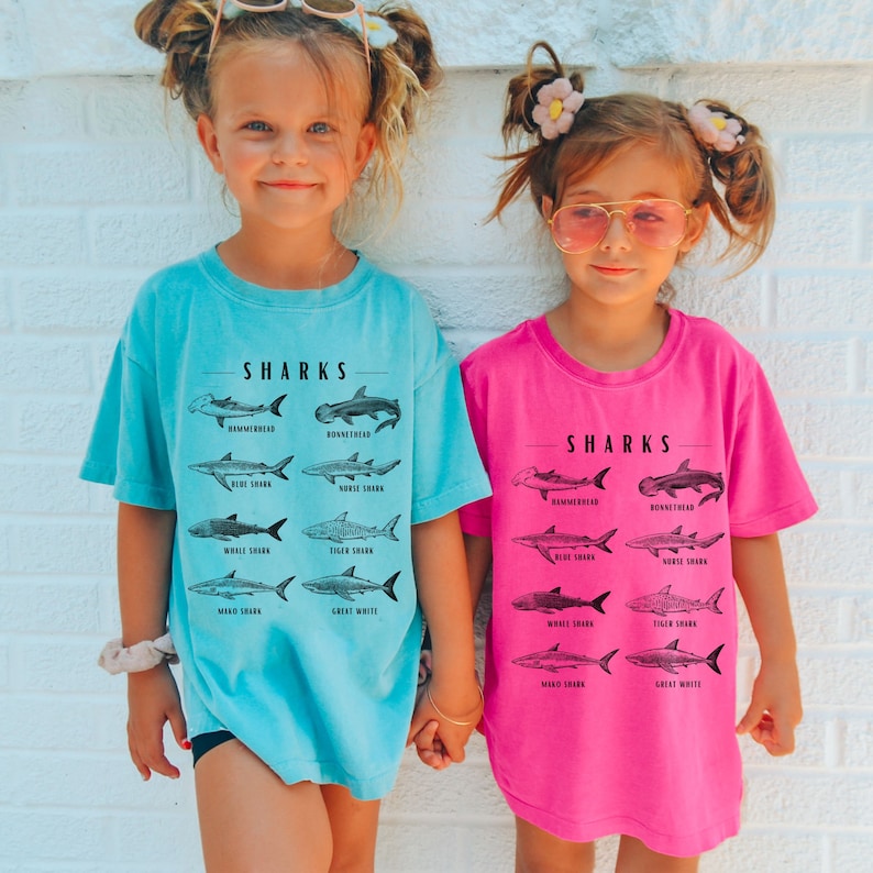 May include: Two children wearing t-shirts with a graphic of different types of sharks and the word "SHARKS" printed on them. The girl on the left is wearing a light blue shirt and the girl on the right is wearing a pink shirt.