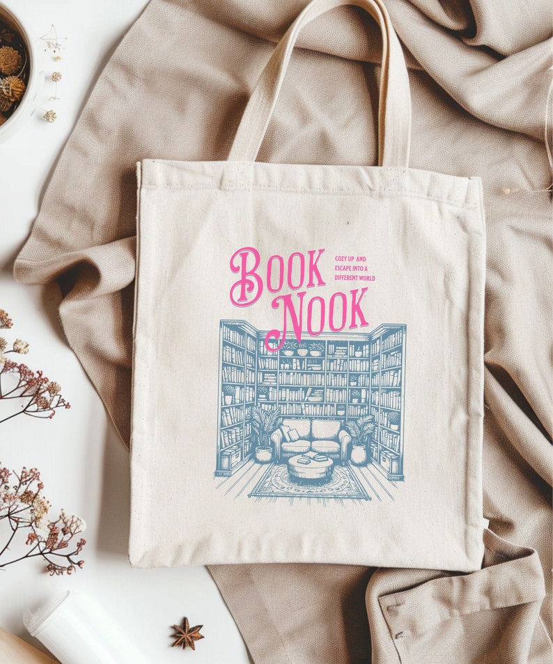 K&ouml;nnte beinhalten: Eine wei&szlig;e Canvas-Tragetasche mit einer blauen und wei&szlig;en Illustration eines B&uuml;cherregals und einer gem&uuml;tlichen Leseecke. Der Text "Book Nook" ist in rosa Schrift mit dem Slogan "Cozy up and escape into a different world".
