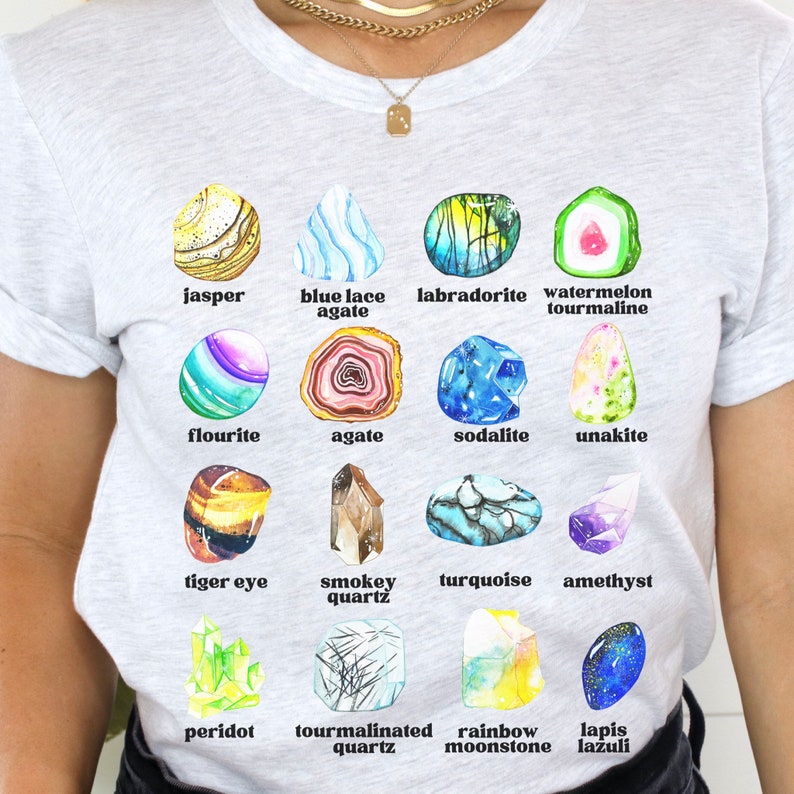 Op de afbeelding: Een wit T-shirt met een kleurrijke illustratie van 14 verschillende soorten edelstenen. Elke edelsteen is gelabeld met zijn naam, waaronder jaspis, blauwe kant-agaten, labradoriet, watermeloen-tourmaline, fluoriet, agaat, sodaliet, unakiet, tijgeroog, rookkwarts, turquoise, amethist, peridoot, tourmalijnkwarts, regenboog-maansteen en lapis lazuli.