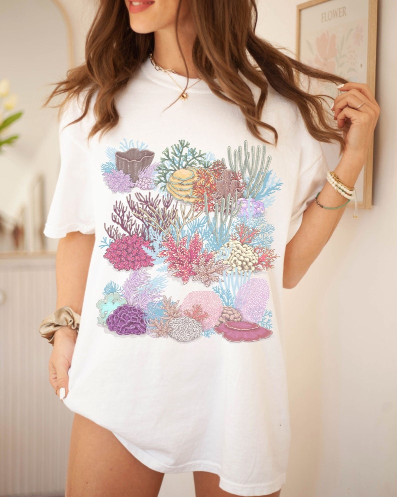 May include: White t-shirt featuring a colourful coral reef design. The design includes various types of coral, sea anemones, and fish in shades of pink, purple, blue, and yellow.