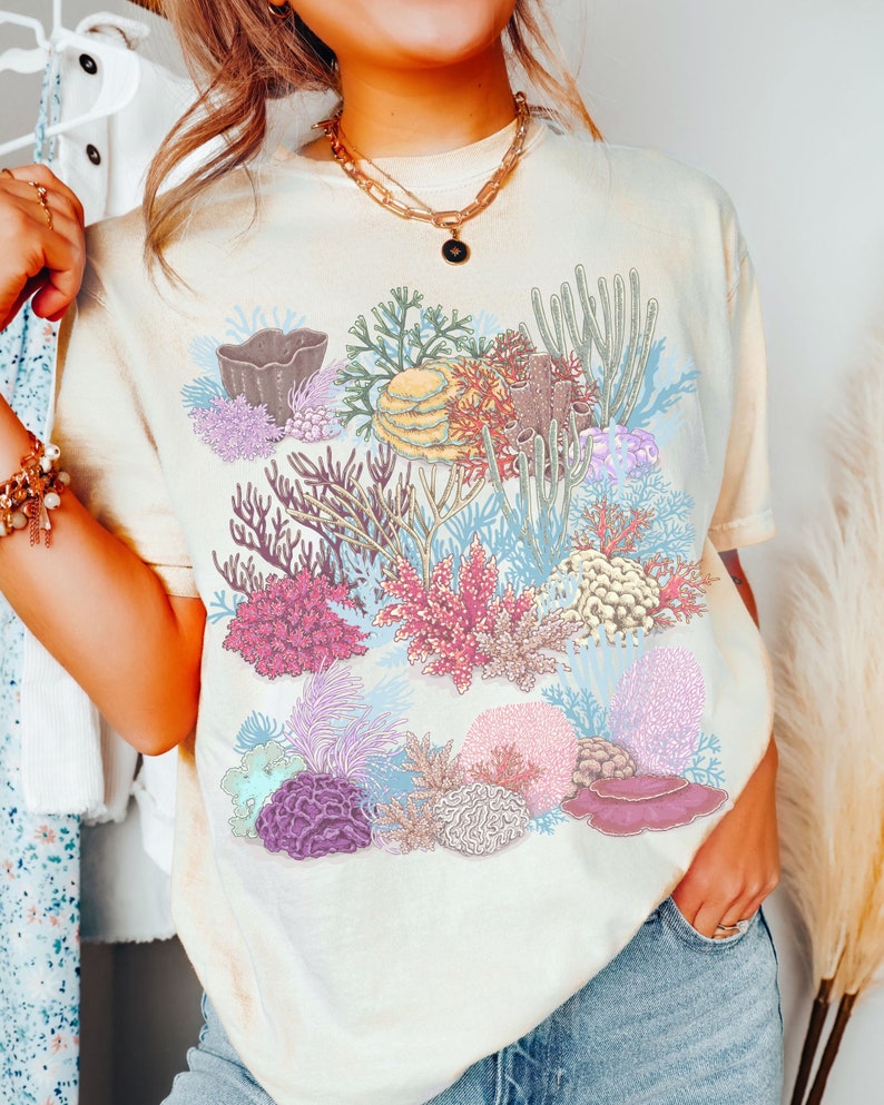 May include: A light yellow t-shirt featuring a colourful coral reef design. The design includes various types of coral, seaweed, and other marine life in shades of pink, purple, blue, and green.