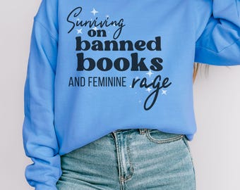 May include: A blue sweatshirt with the text "Surviving on banned books and feminine rage" in black lettering with white stars.