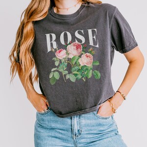 May include: A dark grey t-shirt with a vintage-style floral print of pink roses and green leaves. The word "ROSE" is printed in white above the floral design.