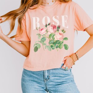 May include: A peach-coloured cropped t-shirt with a white graphic of the word "ROSE" and a vintage illustration of pink roses with green leaves.