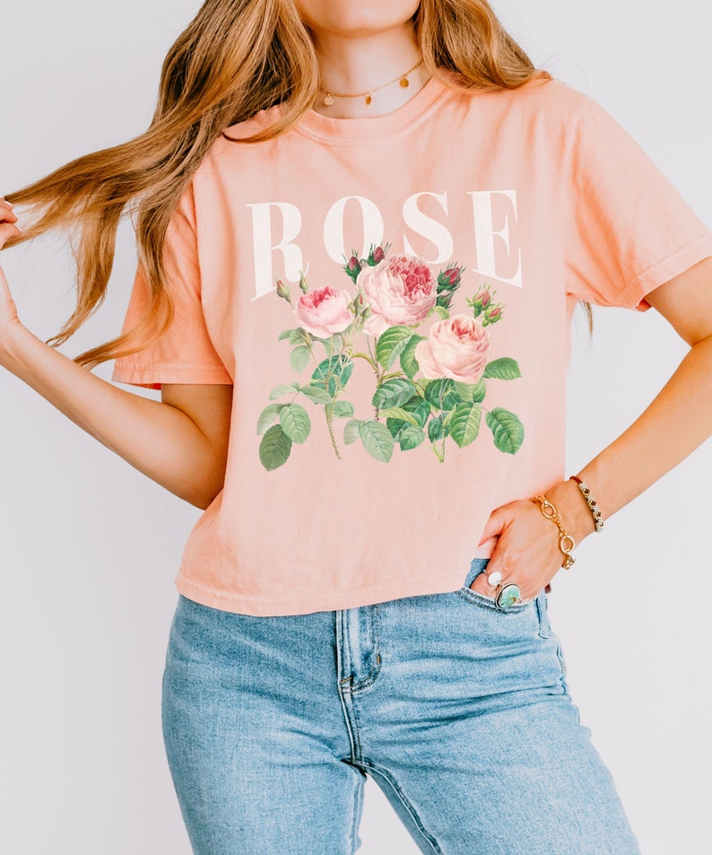 May include: A peach-coloured cropped t-shirt with a white graphic of the word "ROSE" and a vintage illustration of pink roses with green leaves.