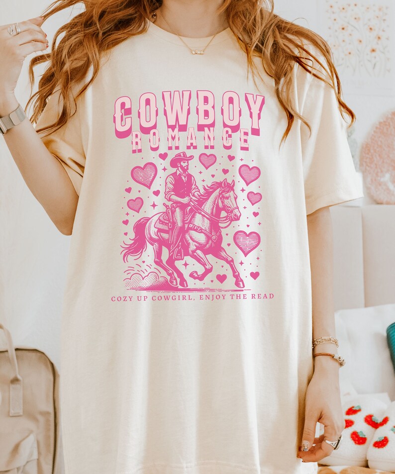 May include: Cream-colored t-shirt with a pink graphic of a cowboy on a horse, surrounded by hearts. The text "COWBOY ROMANCE" is above the image, and "COZY UP COWGIRL, ENJOY THE READ" is below.