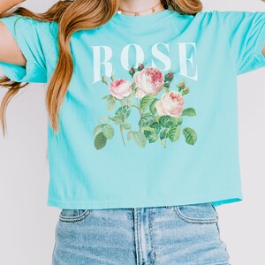 May include: A light blue cropped t-shirt with a white graphic of roses and the word "ROSE" printed on the front.