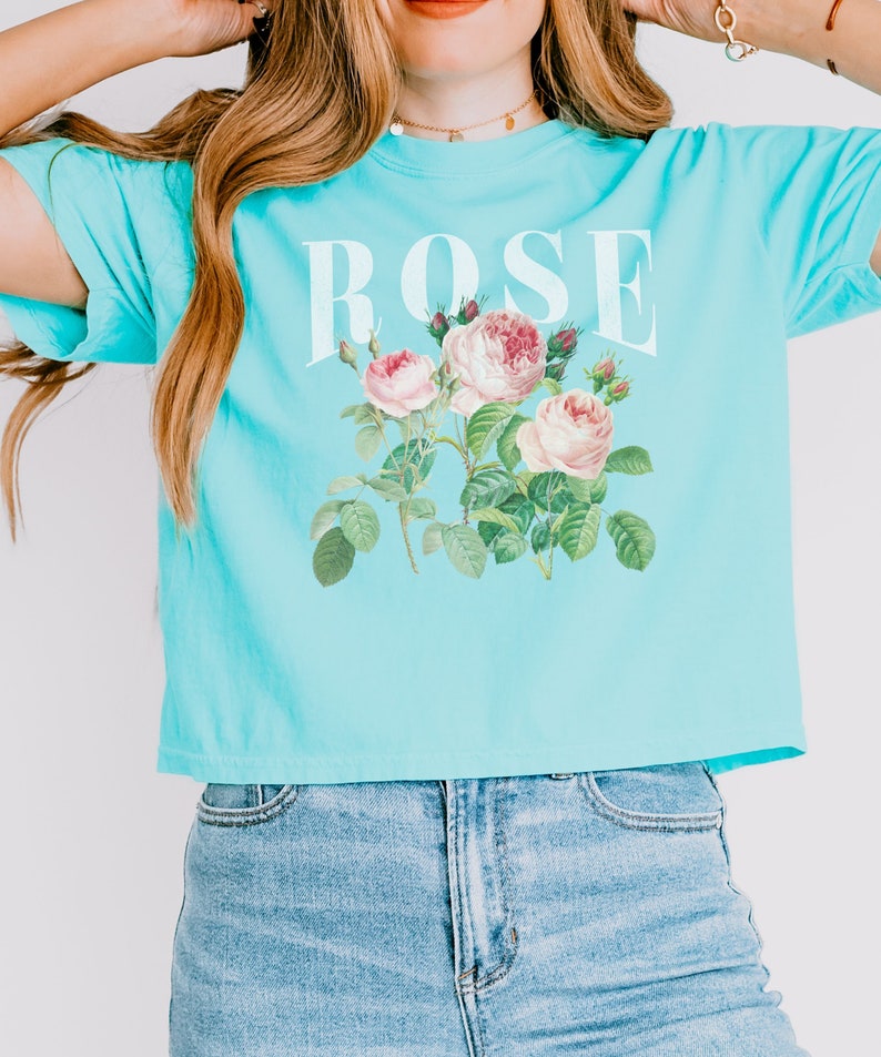 May include: A light blue cropped t-shirt with a white graphic of roses and the word "ROSE" printed on the front.