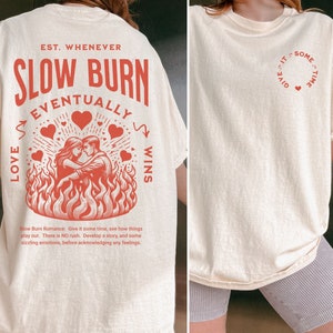 Op de afbeelding: Cr&egrave;mekleurig T-shirt met een rode afbeelding op de rug met de tekst "SLOW BURN EVENTUALLY LOVE WINS" en een illustratie van een stel in vlammen. De voorkant van het shirt heeft een klein rood hartontwerp met de tekst "GIVE IT SOME TIME".