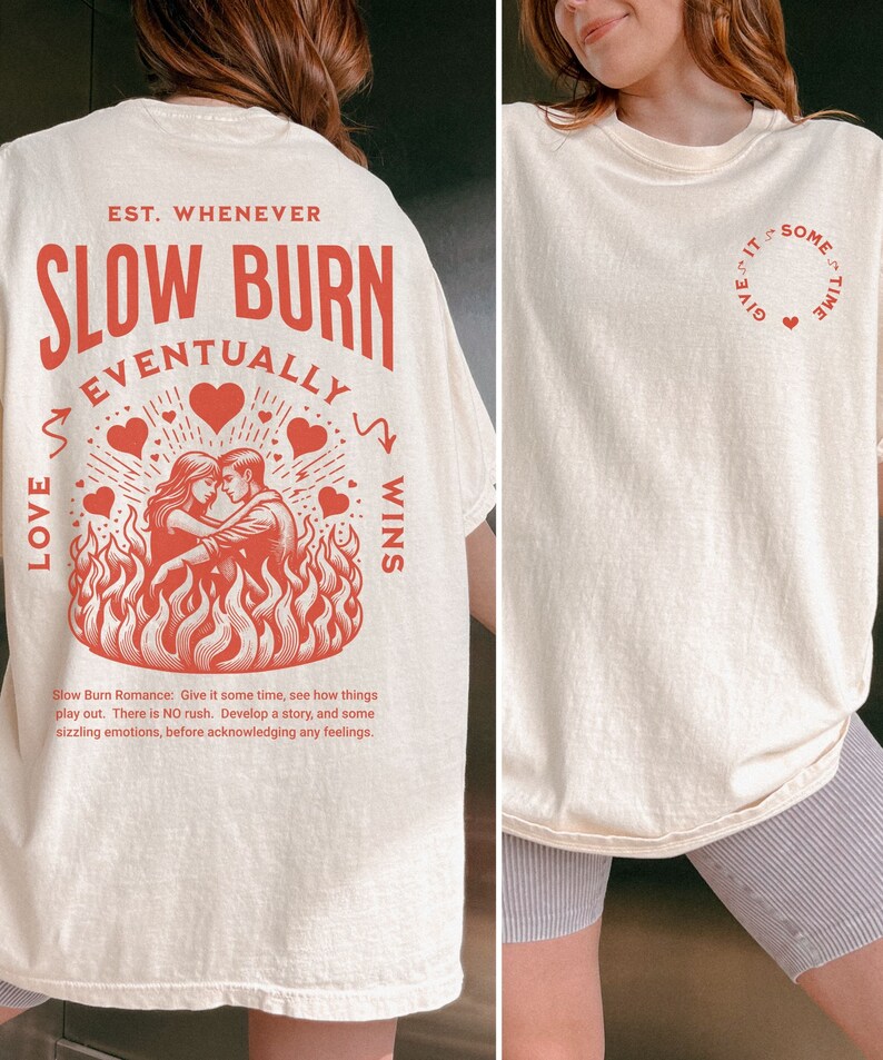 Op de afbeelding: Cr&egrave;mekleurig T-shirt met een rode afbeelding op de rug met de tekst "SLOW BURN EVENTUALLY LOVE WINS" en een illustratie van een stel in vlammen. De voorkant van het shirt heeft een klein rood hartontwerp met de tekst "GIVE IT SOME TIME".