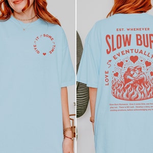 Op de afbeelding: Lichtblauw T-shirt met rode tekst en afbeeldingen. De voorkant heeft een cirkelvormig ontwerp met de tekst "Give it some time" en een hart. De achterkant toont "Slow Burn Eventually Wins" met een stel in vlammen.