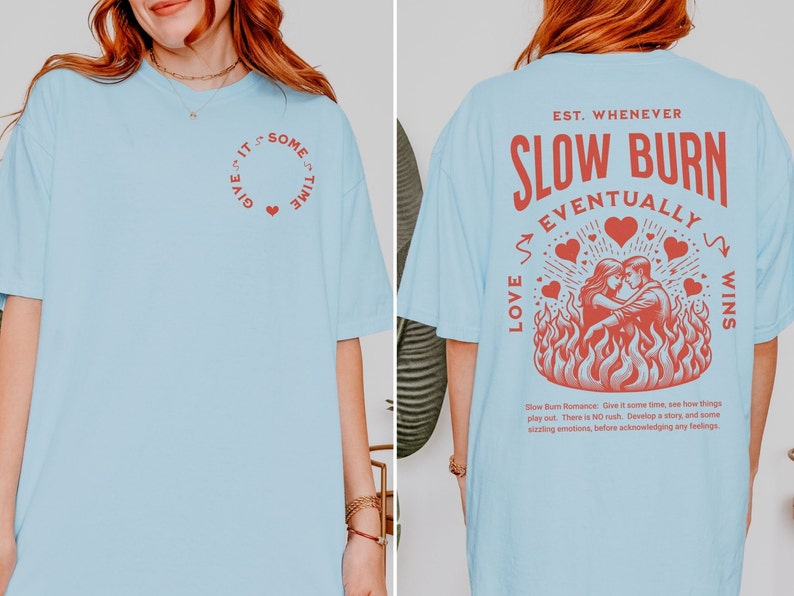 Op de afbeelding: Lichtblauw T-shirt met rode tekst en afbeeldingen. De voorkant heeft een cirkelvormig ontwerp met de tekst "Give it some time" en een hart. De achterkant toont "Slow Burn Eventually Wins" met een stel in vlammen.