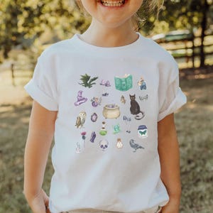 May include: White t-shirt featuring a collection of whimsical watercolor illustrations. The design includes a black cat, a cauldron, an owl, a skull, a spell book, and other magical elements. The shirt is a short-sleeved crew neck.