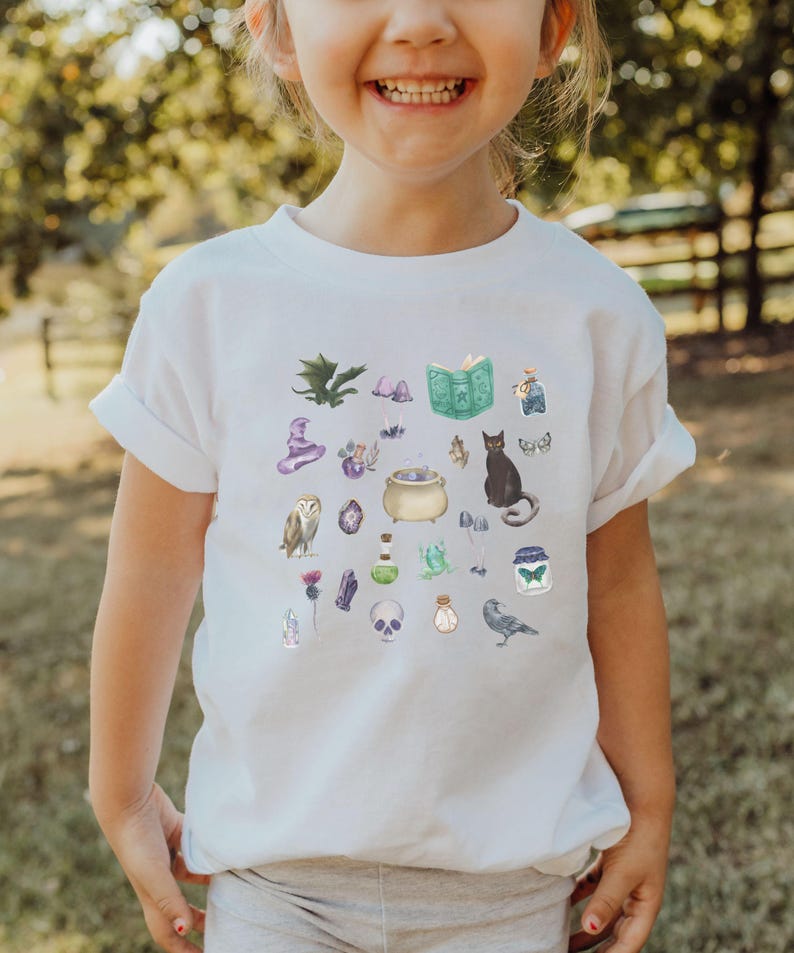 May include: White t-shirt featuring a collection of whimsical watercolor illustrations. The design includes a black cat, a cauldron, an owl, a skull, a spell book, and other magical elements. The shirt is a short-sleeved crew neck.