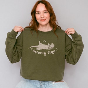 Puede incluir: Una sudadera verde con un gr&aacute;fico de una nutria acostada de espaldas con el texto "otter-ly fine" impreso debajo.
