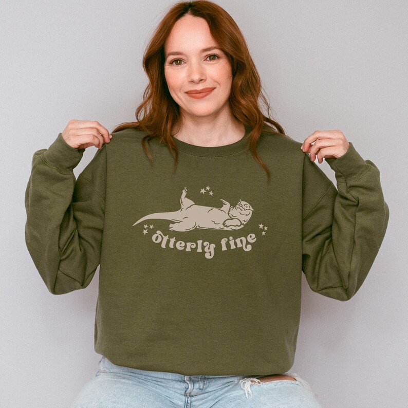Puede incluir: Una sudadera verde con un gr&aacute;fico de una nutria acostada de espaldas con el texto "otter-ly fine" impreso debajo.
