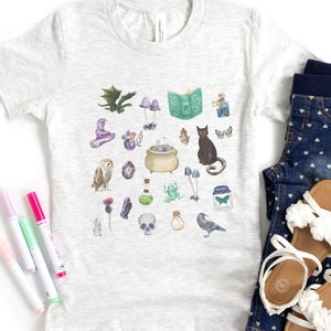 May include: A light gray t-shirt featuring a whimsical watercolor design of magical elements. The design includes a cauldron, owl, black cat, mushrooms, crystals, and a spell book. The shirt is paired with denim shorts and sandals.