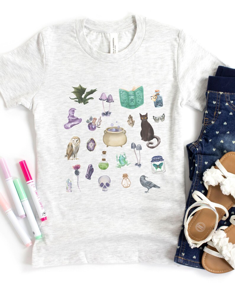 May include: A light gray t-shirt featuring a whimsical watercolor design of magical elements. The design includes a cauldron, owl, black cat, mushrooms, crystals, and a spell book. The shirt is paired with denim shorts and sandals.