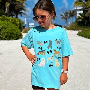 May include: A turquoise blue t-shirt with a graphic print of various dogs wearing black bows. The shirt is made of a soft, comfortable material and has a relaxed fit.