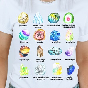Op de afbeelding: Een wit T-shirt met een kleurrijke illustratie van 12 verschillende soorten edelstenen. Elke edelsteen is gelabeld met zijn naam, waaronder jaspis, blauwe kant-agaten, labradoriet, watermeloen-tourmaline, fluoriet, agaat, sodaliet, unakiet, tijgeroog, rookkwarts, turquoise, amethist, peridoot, tourmalijnkwarts, regenboog-maansteen en lapis lazuli.