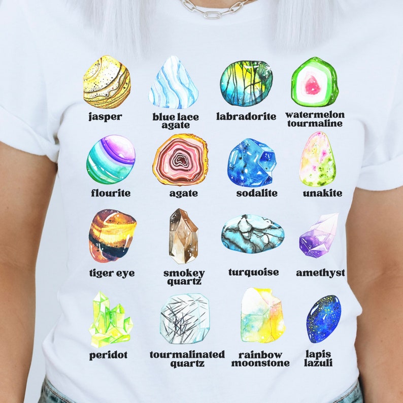 Op de afbeelding: Een wit T-shirt met een kleurrijke illustratie van 12 verschillende soorten edelstenen. Elke edelsteen is gelabeld met zijn naam, waaronder jaspis, blauwe kant-agaten, labradoriet, watermeloen-tourmaline, fluoriet, agaat, sodaliet, unakiet, tijgeroog, rookkwarts, turquoise, amethist, peridoot, tourmalijnkwarts, regenboog-maansteen en lapis lazuli.