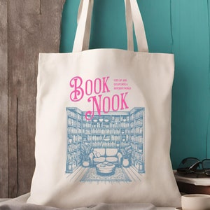 K&ouml;nnte beinhalten: Eine wei&szlig;e Canvas-Tragetasche mit einem blauen und rosafarbenen Design. Das Design zeigt ein B&uuml;cherregal mit einer Couch und einem Couchtisch. Der Text "BOOK NOOK" ist in Rosa geschrieben, der Text "COZY UP AND ESCAPE INTO A DIFFERENT WORLD" in kleinerer blauer Schrift.