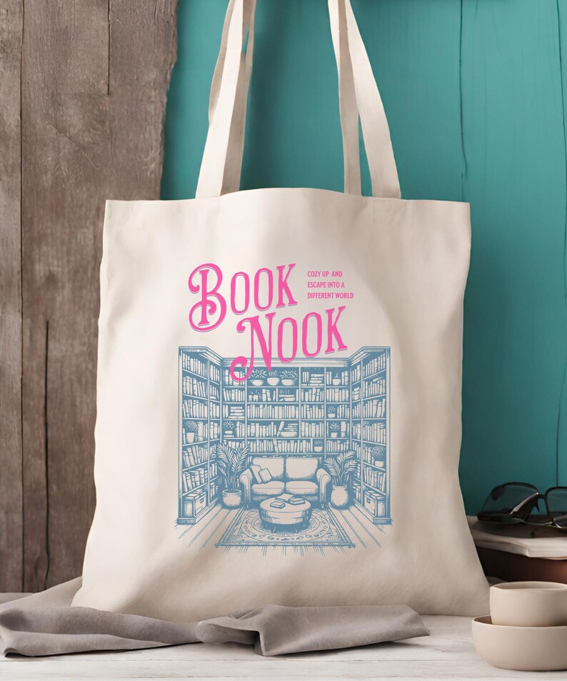 K&ouml;nnte beinhalten: Eine wei&szlig;e Canvas-Tragetasche mit einem blauen und rosafarbenen Design. Das Design zeigt ein B&uuml;cherregal mit einer Couch und einem Couchtisch. Der Text "BOOK NOOK" ist in Rosa geschrieben, der Text "COZY UP AND ESCAPE INTO A DIFFERENT WORLD" in kleinerer blauer Schrift.